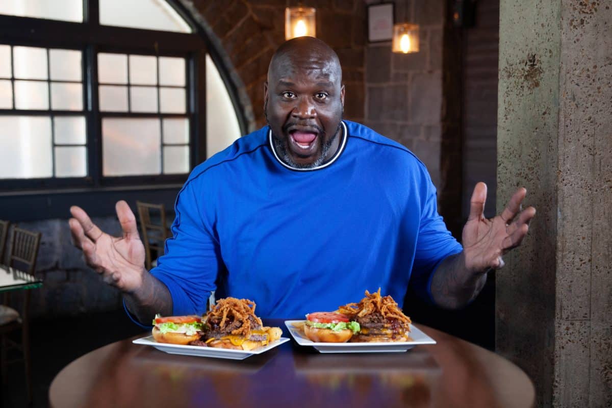 Shaq sits at a table in front of two plates of sandwiches.